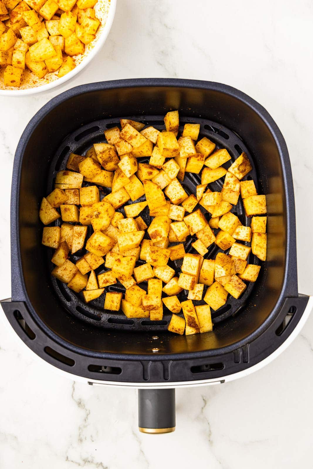 overhead view of black air fryer containing seasoned diced potatoes, with white bowl of diced potatoes in background