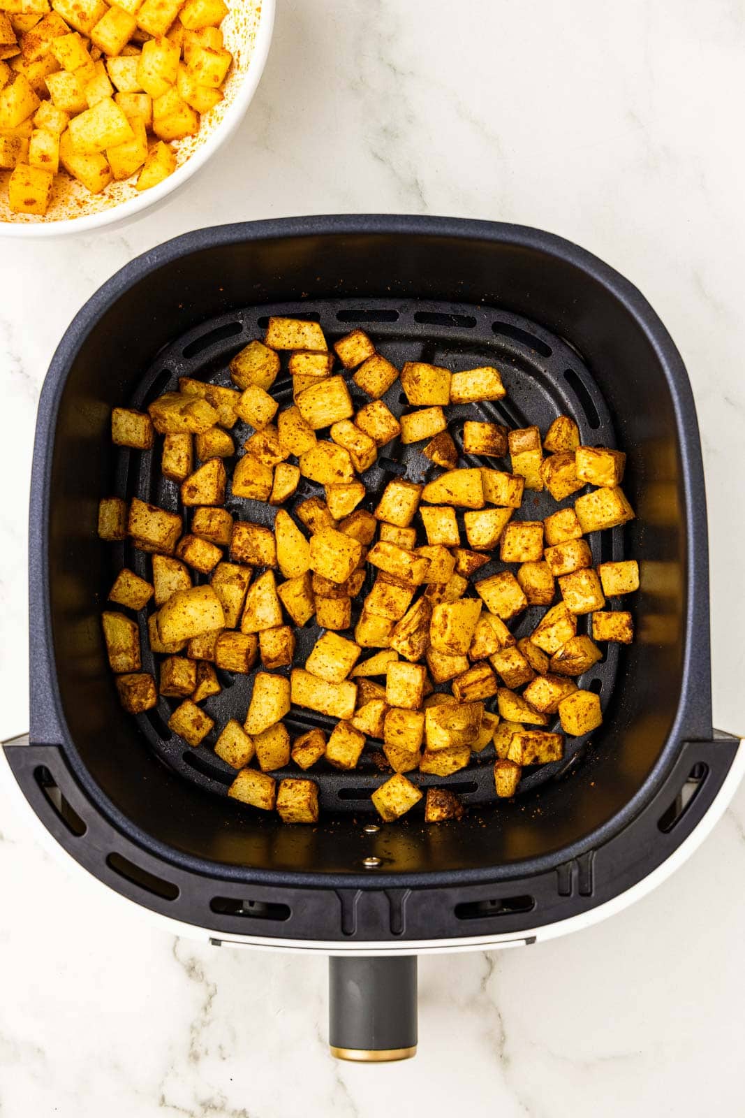 overhead view of black air fryer containing air fried seasoned diced potatoes, with white bowl of diced potatoes in background