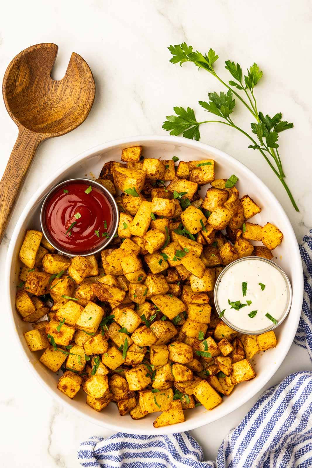 overhead view of white bowl containing air fried diced potatoes with containers of ketchup and ranch, surrounded by wooden spoon, parsley sprig and white and blue striped dish towel