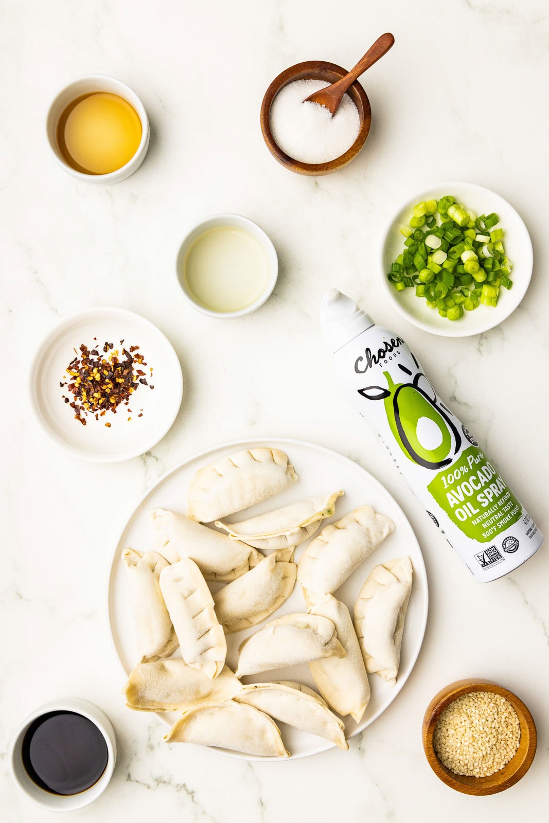 ingredients to include white plate containing uncooked pot stickers, can of avocado oil spray, containers of sliced green onions, sesame seeds, soy sauce, rice vinegar, sesame oil, chili flakes, sugar