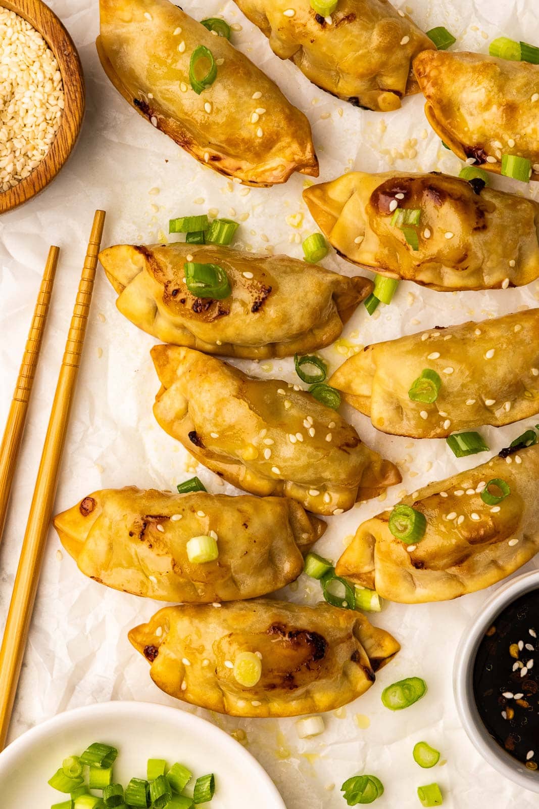 overhead view of 10 air fried pot stickers sprinkled with sesame seeds with white bowl of dipping sauce, wooden bowl of sesame seeds white bowl of green onions and a set of chopsticks on parchment paper