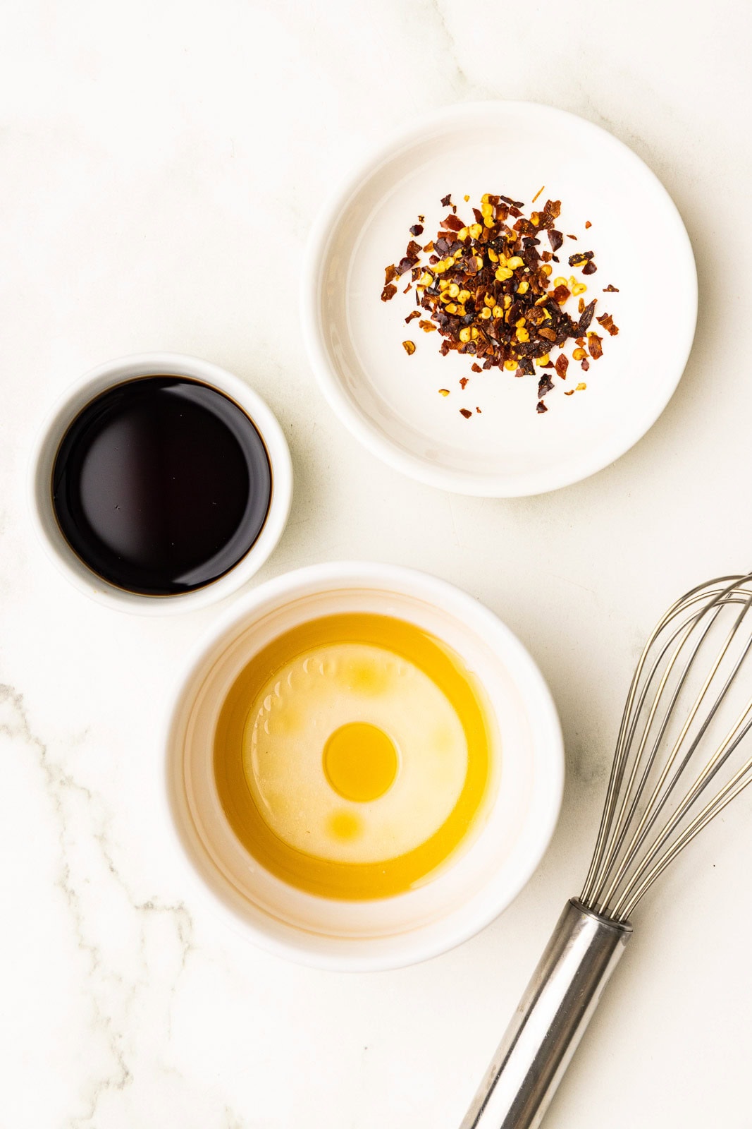 overhead view of white bowls containing soy sauce, chili flakes and sesame oil with silver whisk on side