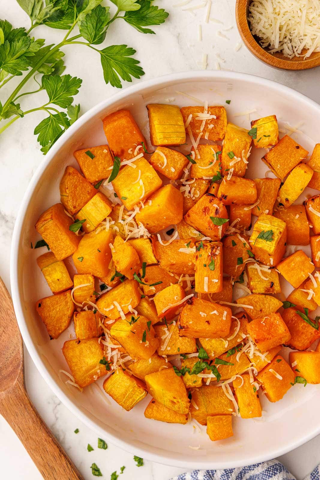 white plate of air fried butternut squash sprinkled with parsley and parmesan cheese, with parsley sprig and parmesan cheese bowl on side