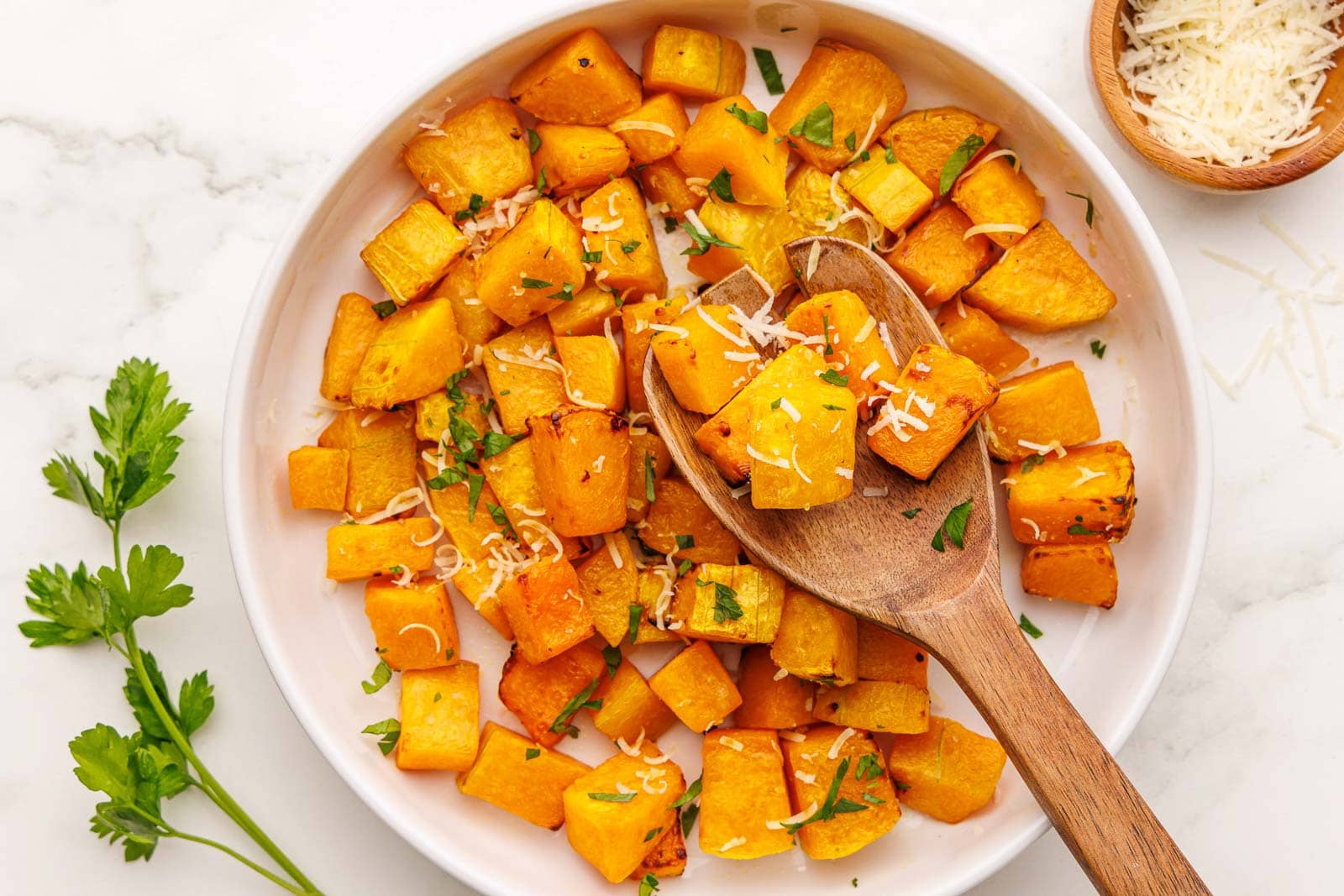 overhead view of plate of air fried butternut squash with large wooden spoon, surrounded by parsley sprig and wooden bowl of parmesan cheese