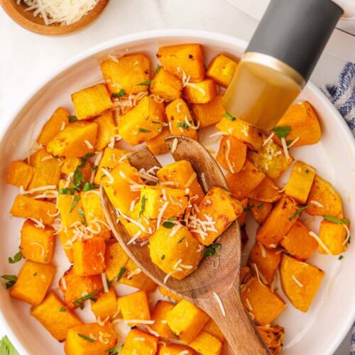 overhead view of plate of air fried butternut squash with large wooden spoon, surrounded by parsley sprig, wooden bowl of parmesan cheese and air fryer