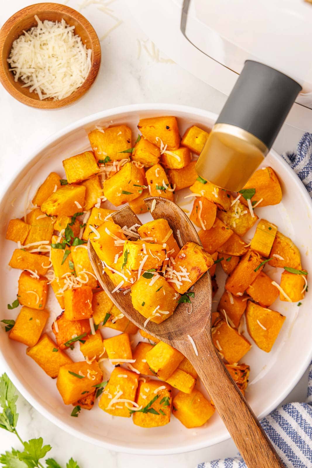 overhead view of plate of air fried butternut squash with large wooden spoon, surrounded by parsley sprig, wooden bowl of parmesan cheese and air fryer