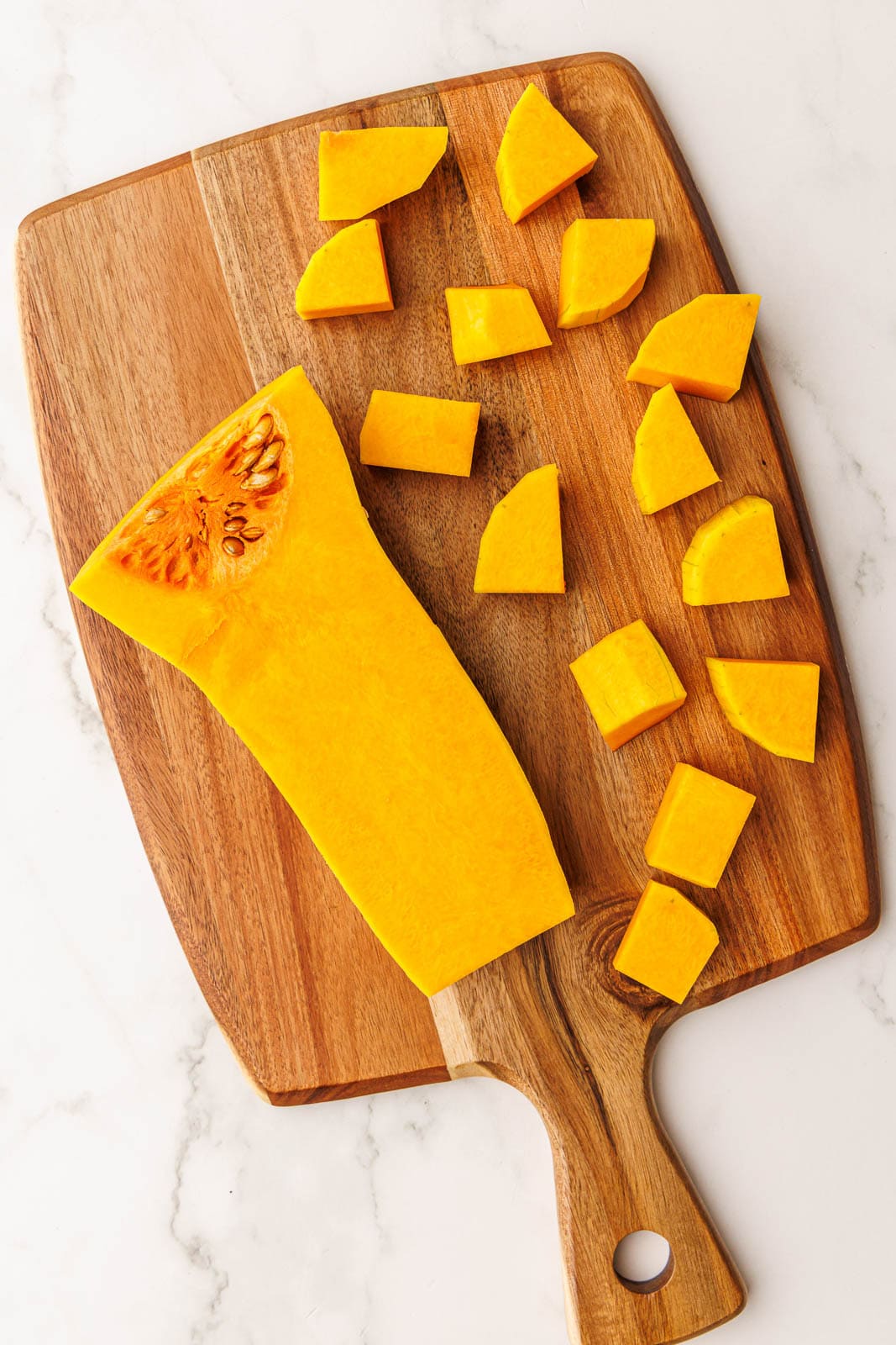 wooden cutting board with one half peeled butternut squash with cubes alongside