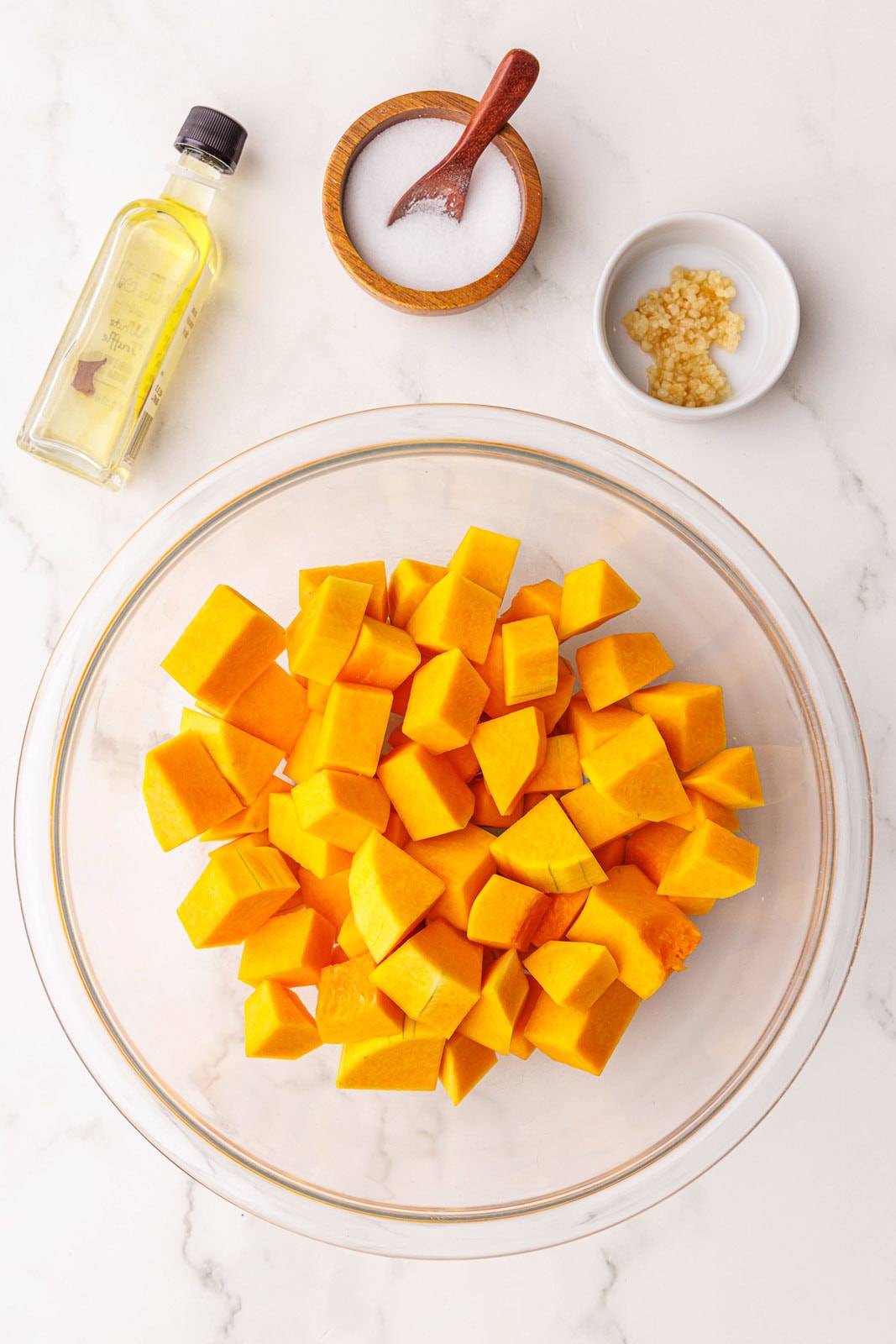 glass bowl filled with squash cubes surrounded by bottle of olive oil, minced garlic and wooden bowl and spoon of salt