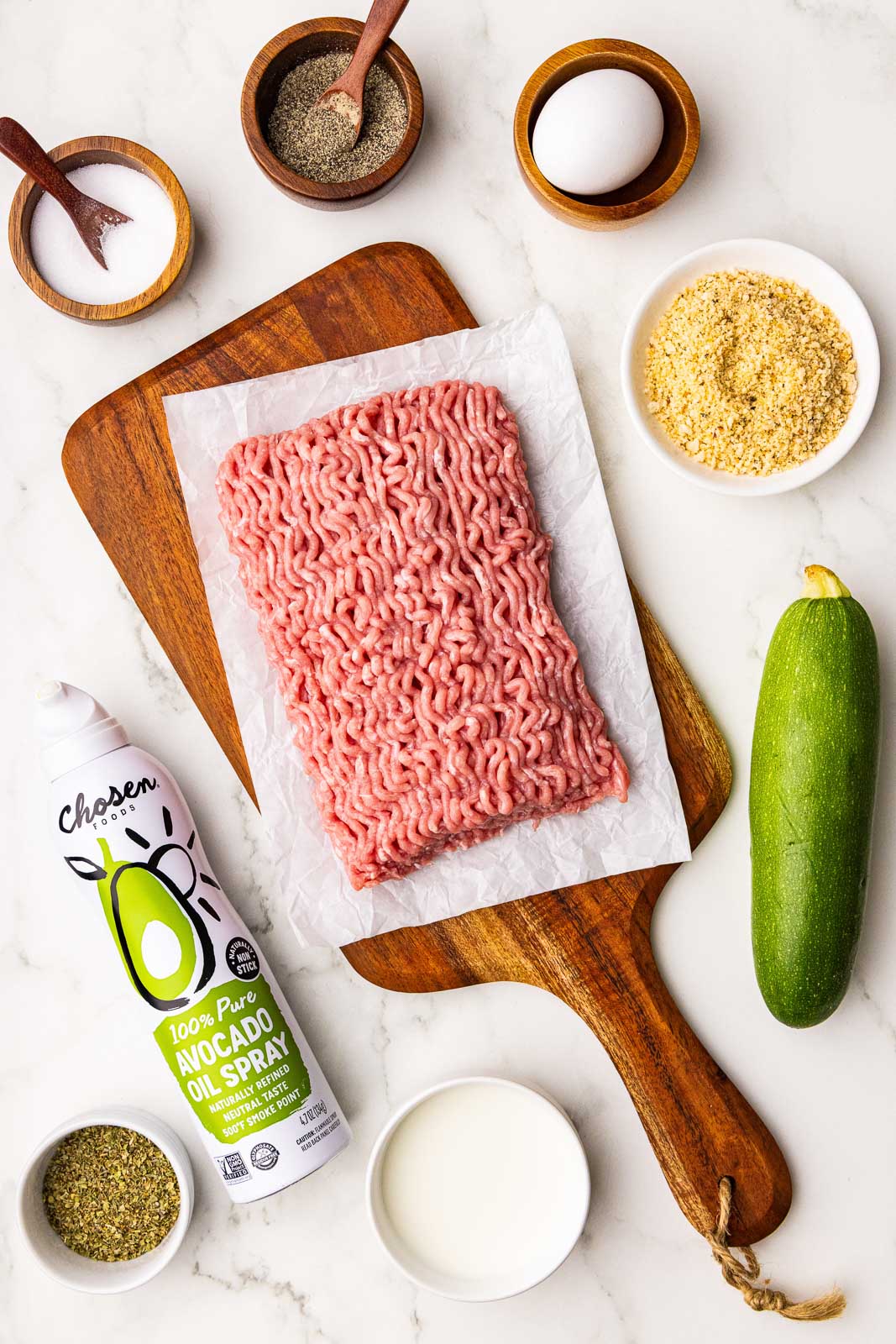 turkey meatball ingredients to include ground turkey on parchment lined wooden cutting board, wooden bowls with spoons of salt and pepper, one egg, bowl of panko breadcrumbs, one zucchini, small bowl of milk, avocado oil spray and bowl of seasoning