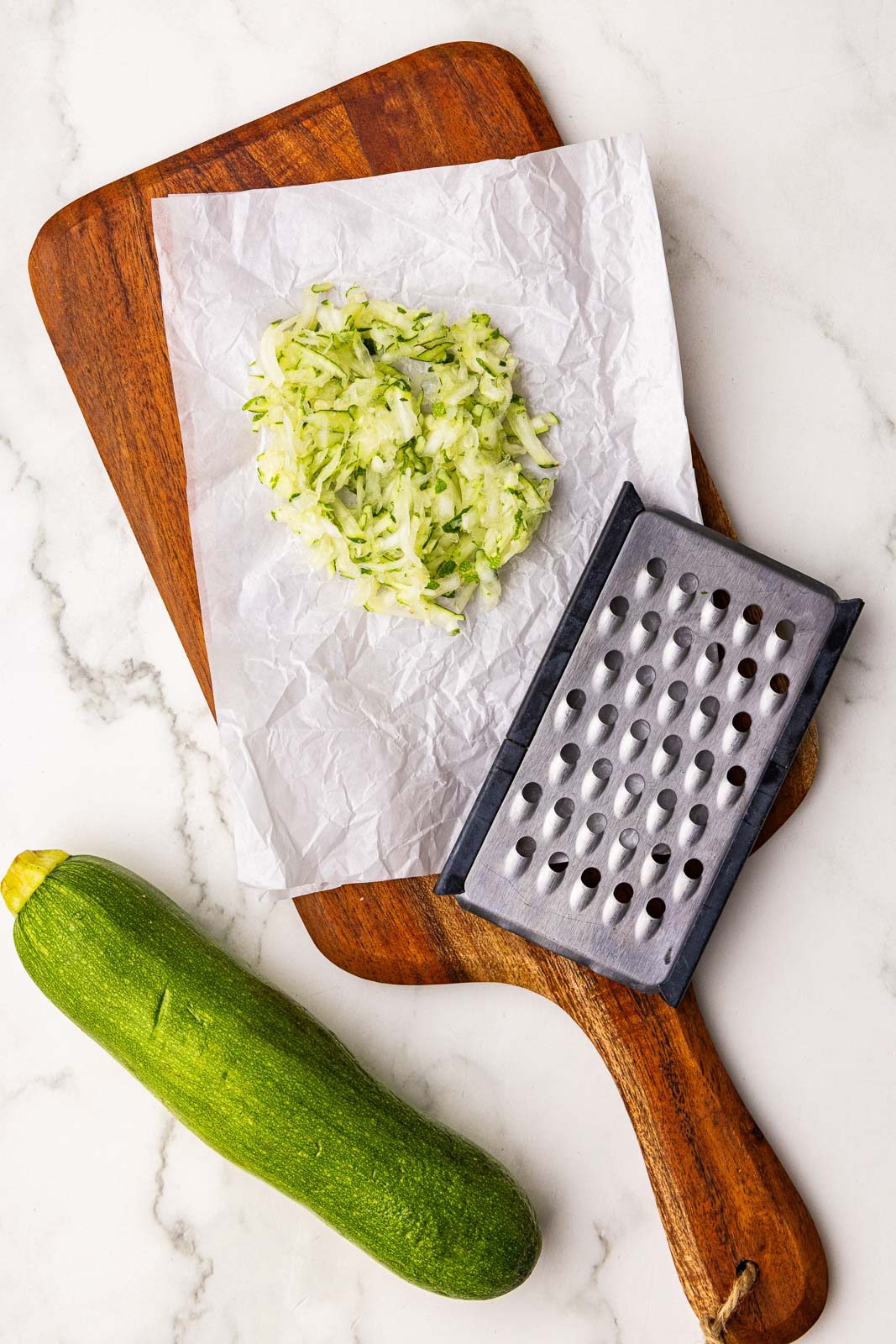 parchment lined wooden cutting board with grated zucchini, grater and zucchini on side