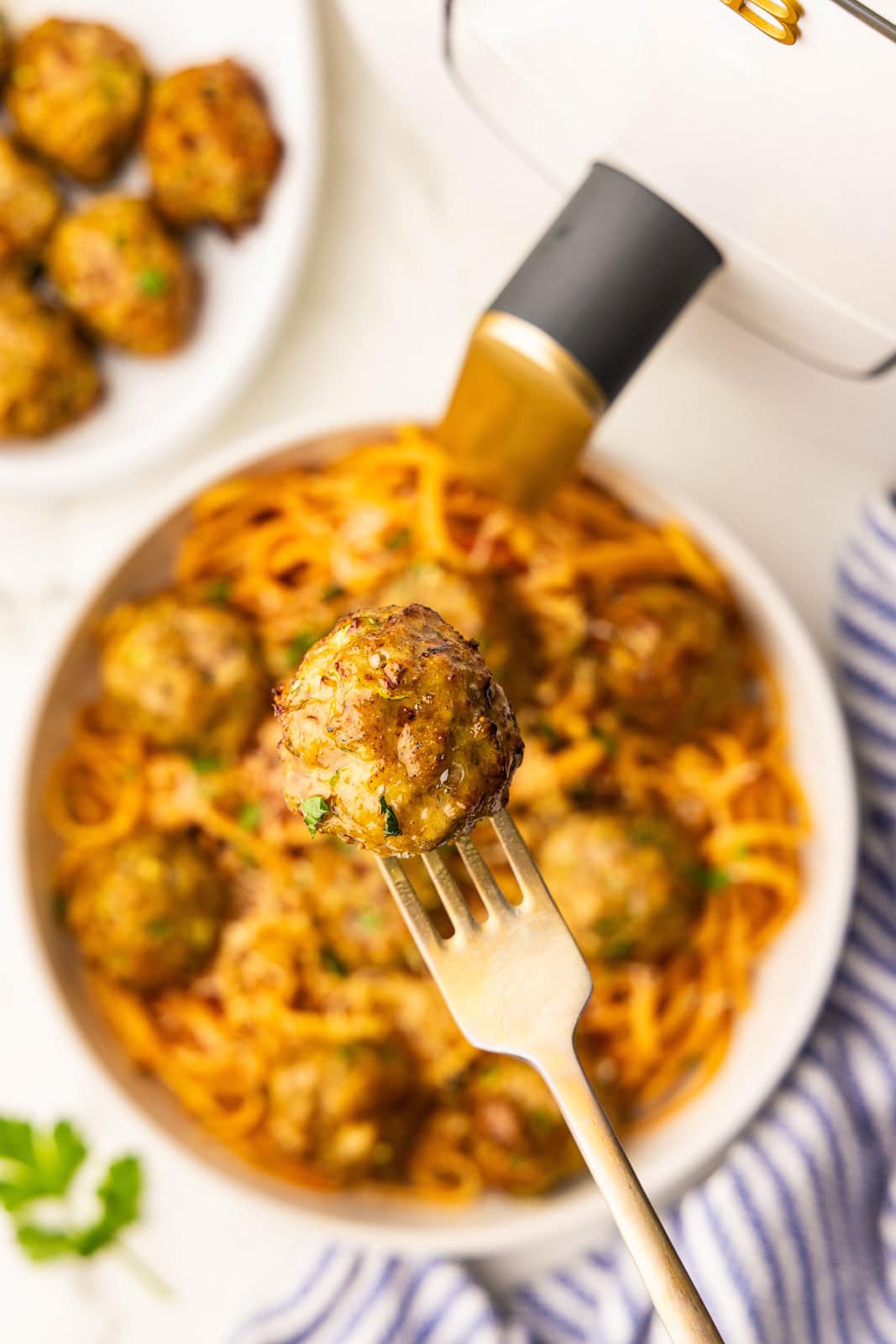 closeup view of fork containing a turkey meatball, with air fryer, plate of turkey meatballs and pasta and platter of turkey meatballs in background