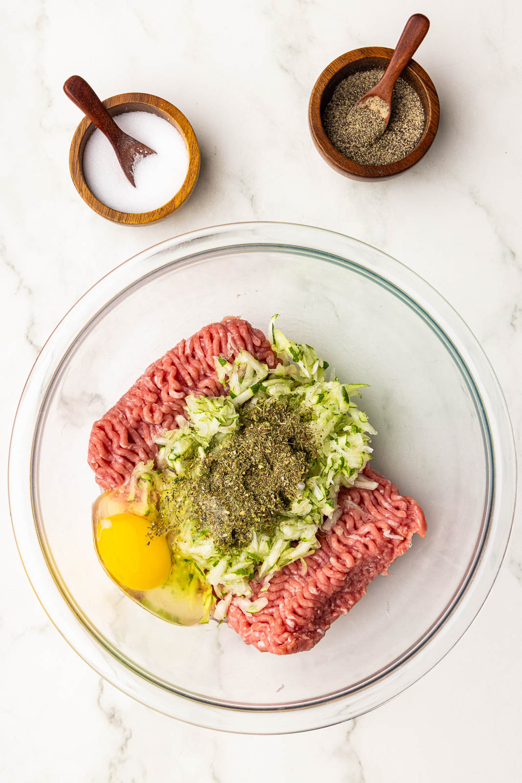 glass bowl with ground turkey, egg, zucchini and seasoning, with wooden bowls and spoons of salt and pepper on side