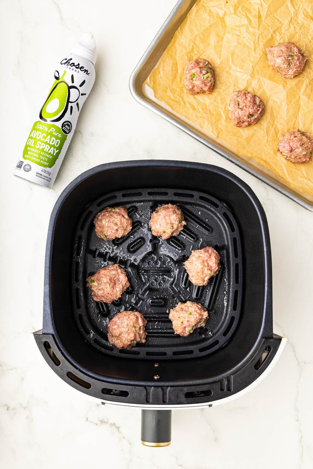 overhead view of black air fryer with 6 meatballs, can of avocado spray and parchment lined baking sheet with meatballs in background
