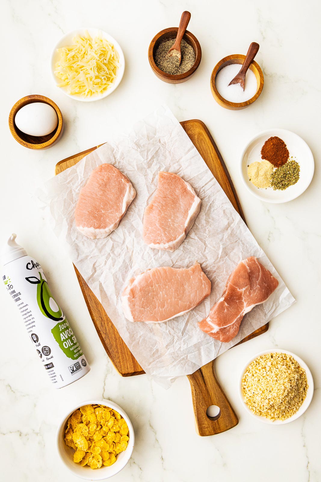 breaded pork chops ingredients to include parchment lined wooden cutting board with flour pork chops surrounded by bowls of corn flakes, panko breadcrumbs, seasoning, salt and pepper, parmesan cheese, one egg, and can of avocado oil spray