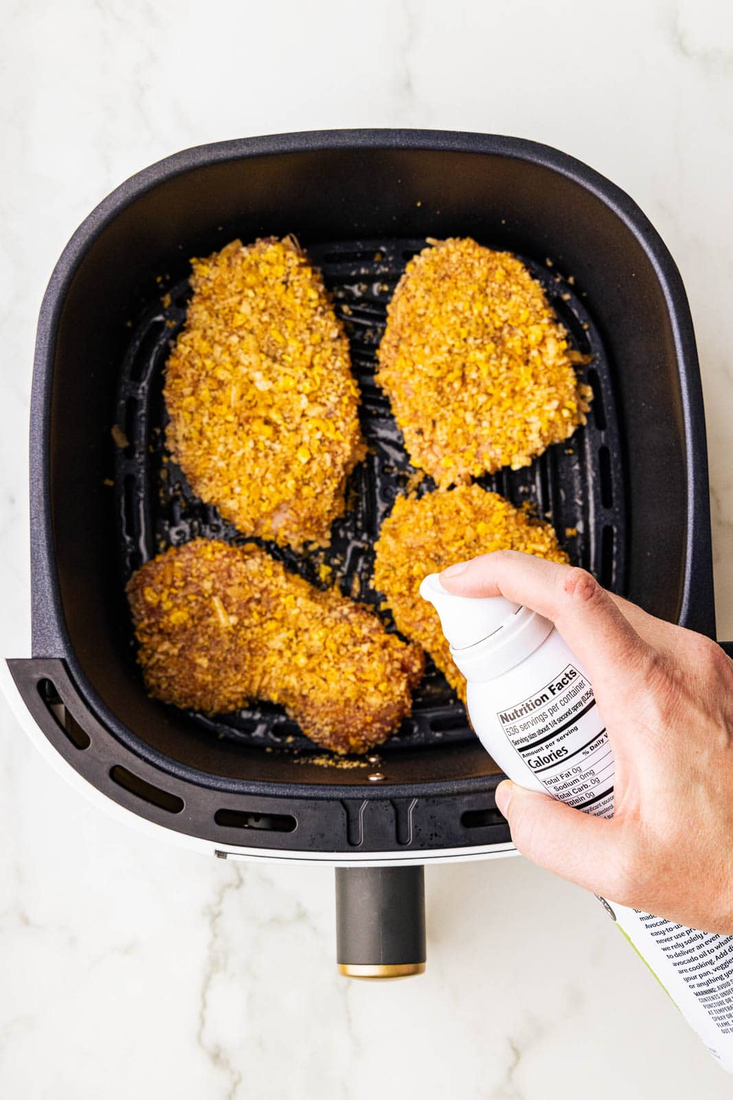 black air fryer with four coated pork chops and hand holding avocado oil spray