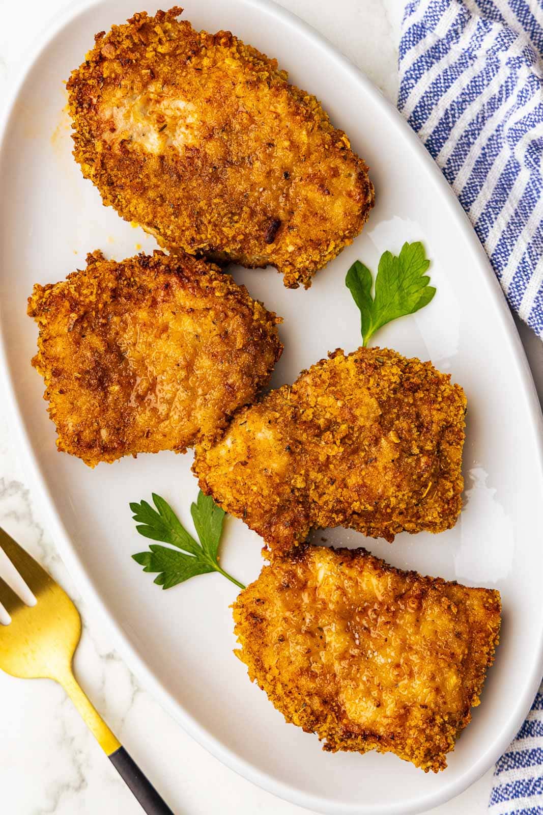 white platter containing four air fried pork chops with gold fork and blue and white dish towel on side