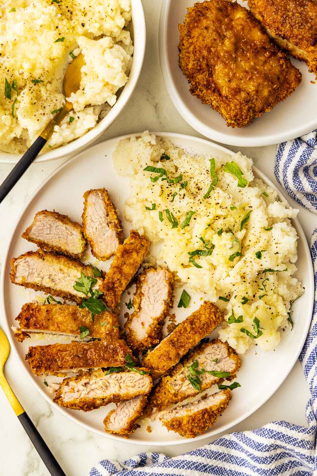 overhead view of dinner plate containing sliced pork chop with mashed potatoes, surrounded by platter of pork chops and bowl of mashed potatoes, along with blue and white striped dish towel and gold fork