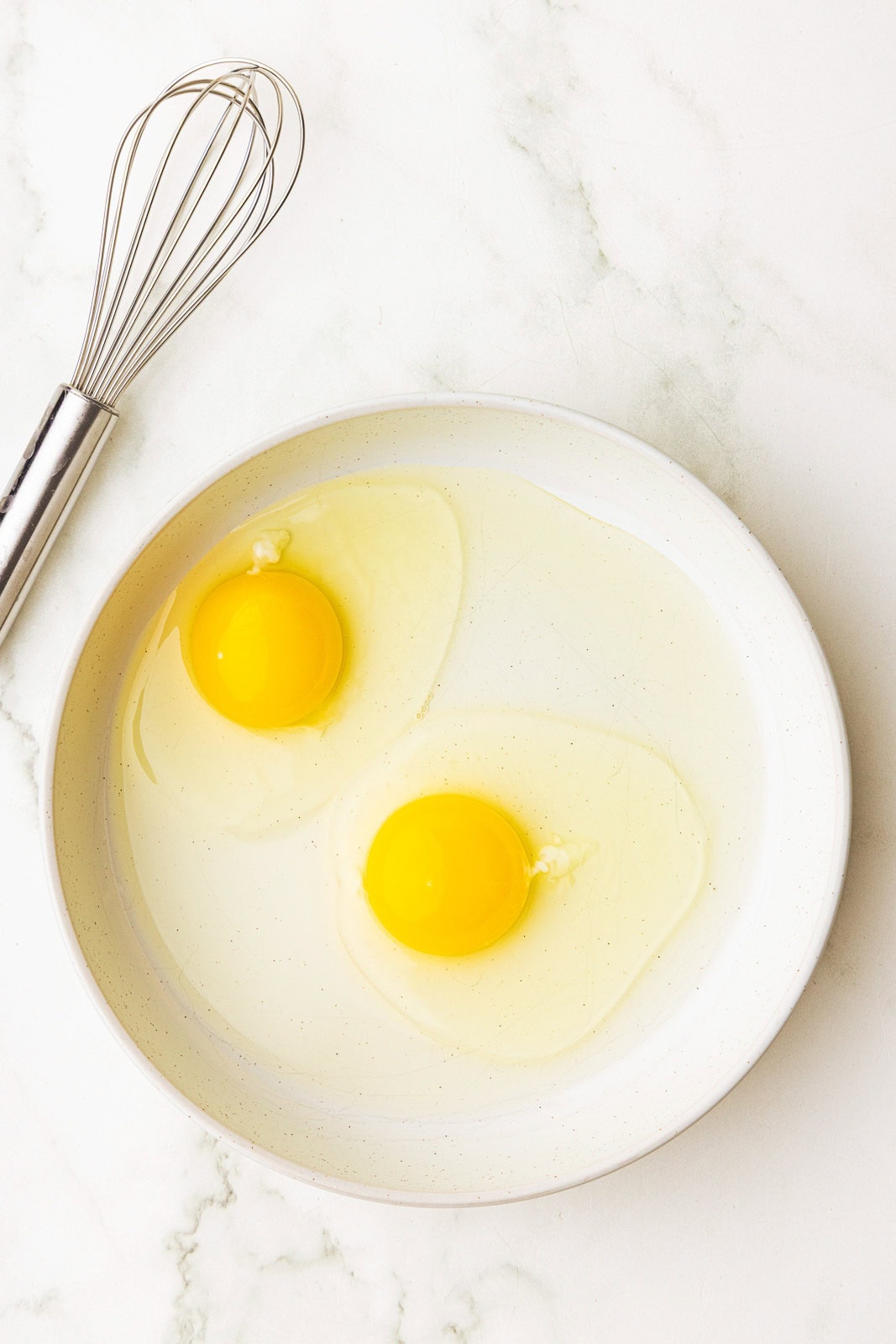 white bowl with two cracked eggs and wire whisk on side