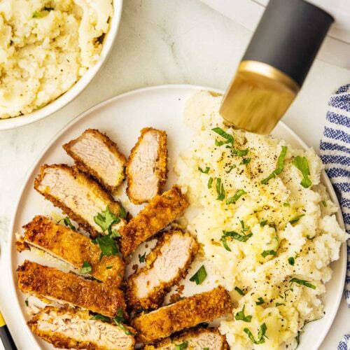 overhead view of dinner plate containing sliced pork chop with mashed potatoes, surrounded by bowl of mashed potatoes and white air fryer, along with blue and white striped dish towel and fork
