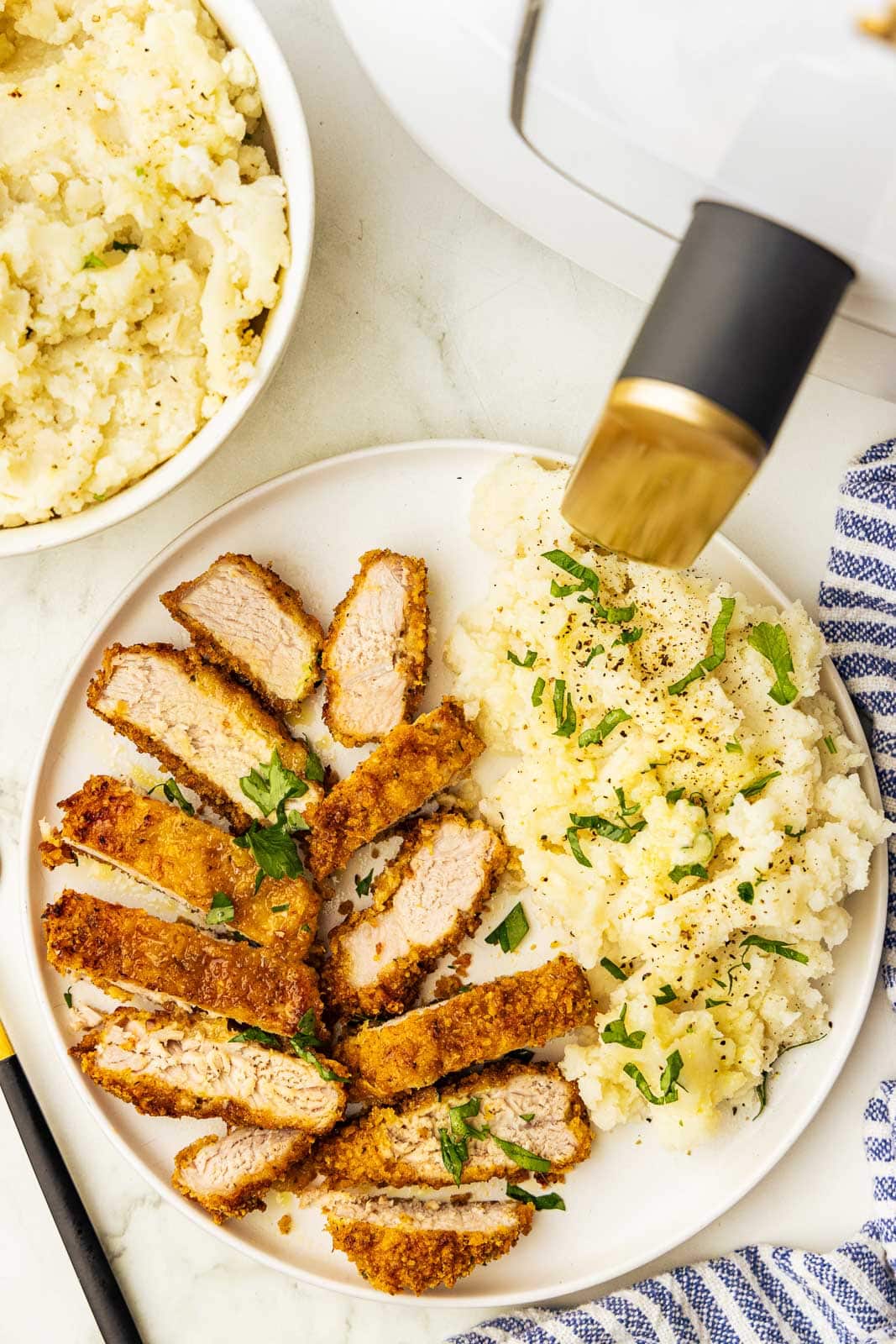 overhead view of dinner plate containing sliced pork chop with mashed potatoes, surrounded by bowl of mashed potatoes and white air fryer, along with blue and white striped dish towel and fork
