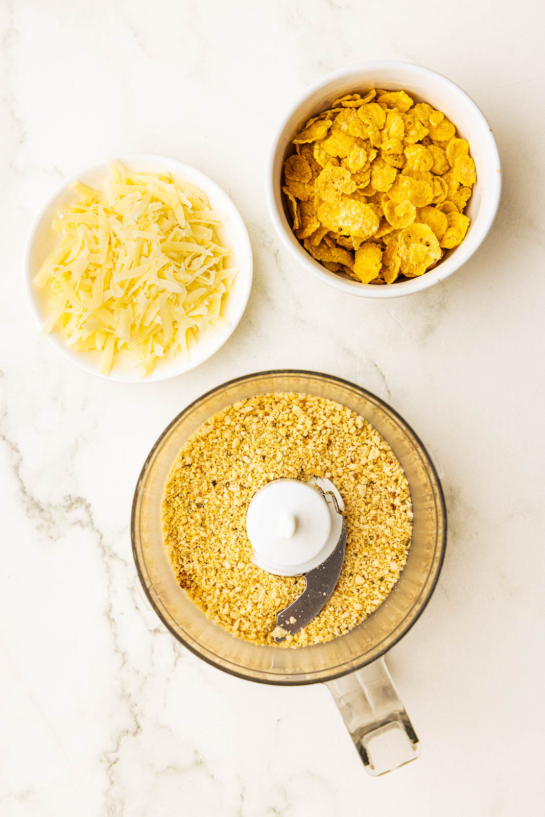 small food processor with bread crumbs, surrounded by bowls of parmesan cheese and corn flakes