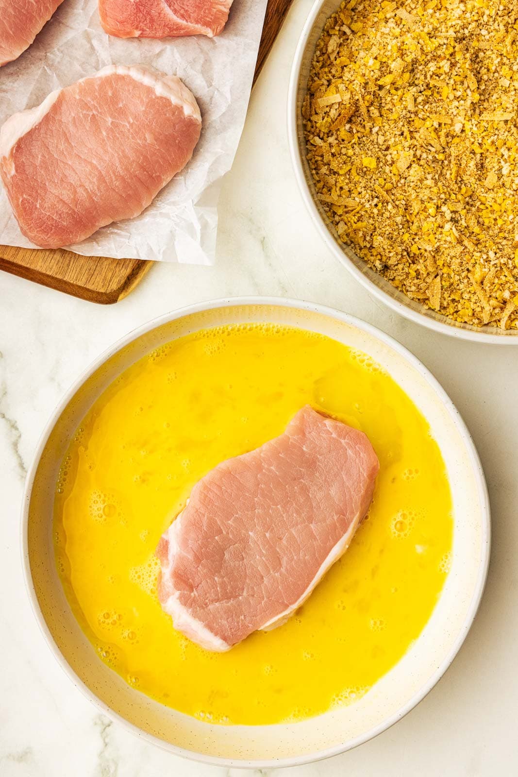 beaten egg mixture with pork chop, with bread crumbs dry mixture and wooden cutting board with pork chops on side