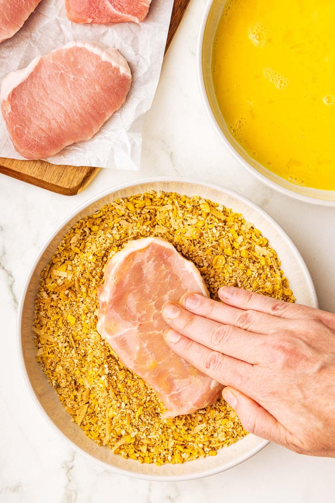 hand inserting pork chop into dry bread mixture, with beaten egg bowl and wooden cutting board with pork chops on side