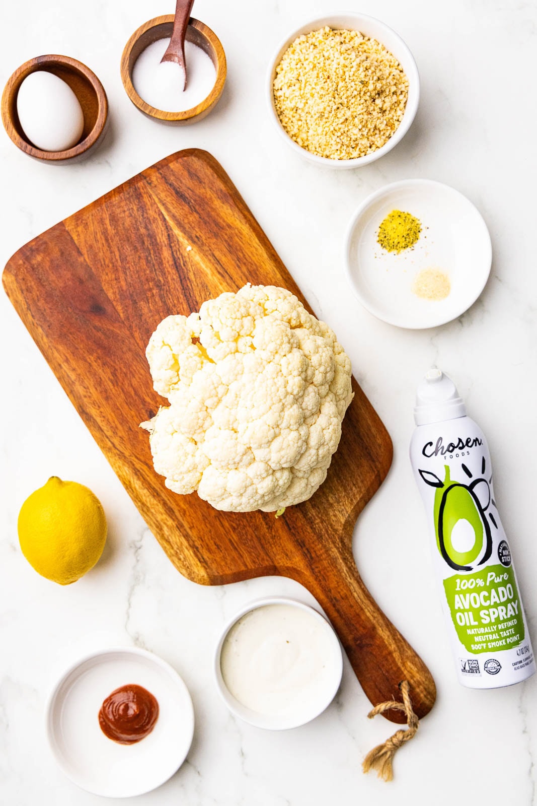 ingredients to include cutting board with head of cauliflower, surrounded by wooden bowls and spoons of salt and egg, with white bowls of panko breadcrumbs, lemon pepper seasoning, garlic powder, ranch dressing, sriracha, can of avocado oil spray, and one lemon