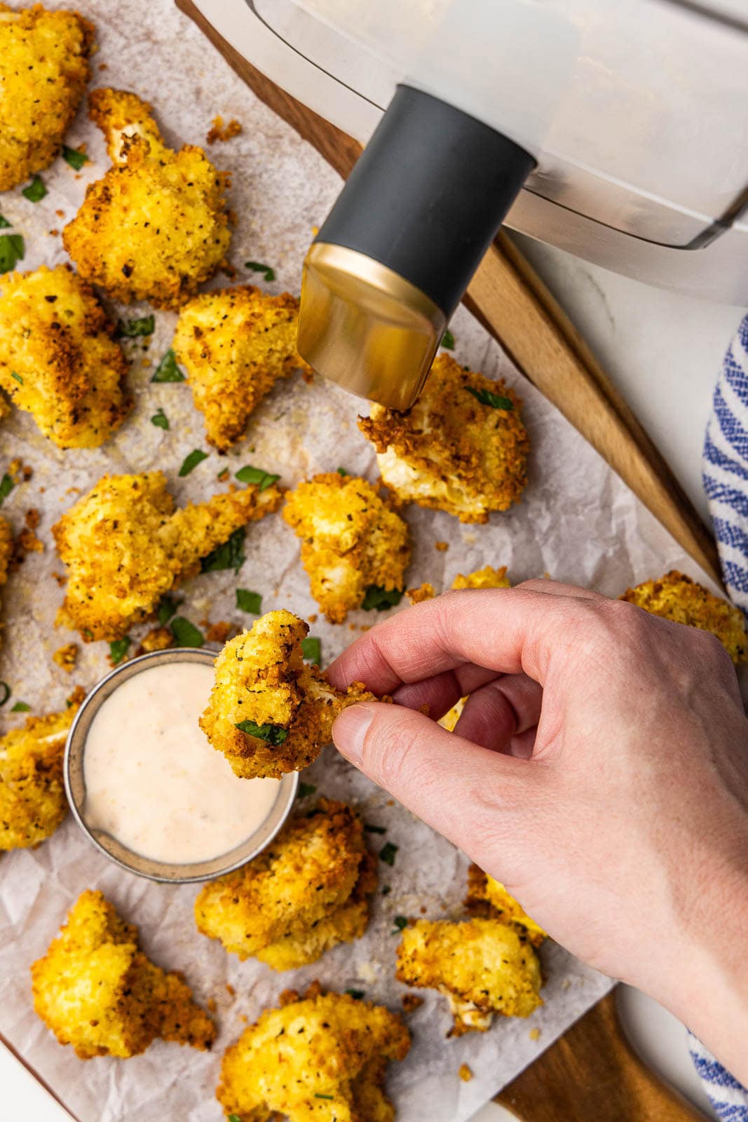 I heard view of parchment lined cooking sheet with cauliflower, with hand inserting one floret into Srirach ranch dip, with white air fryer on side