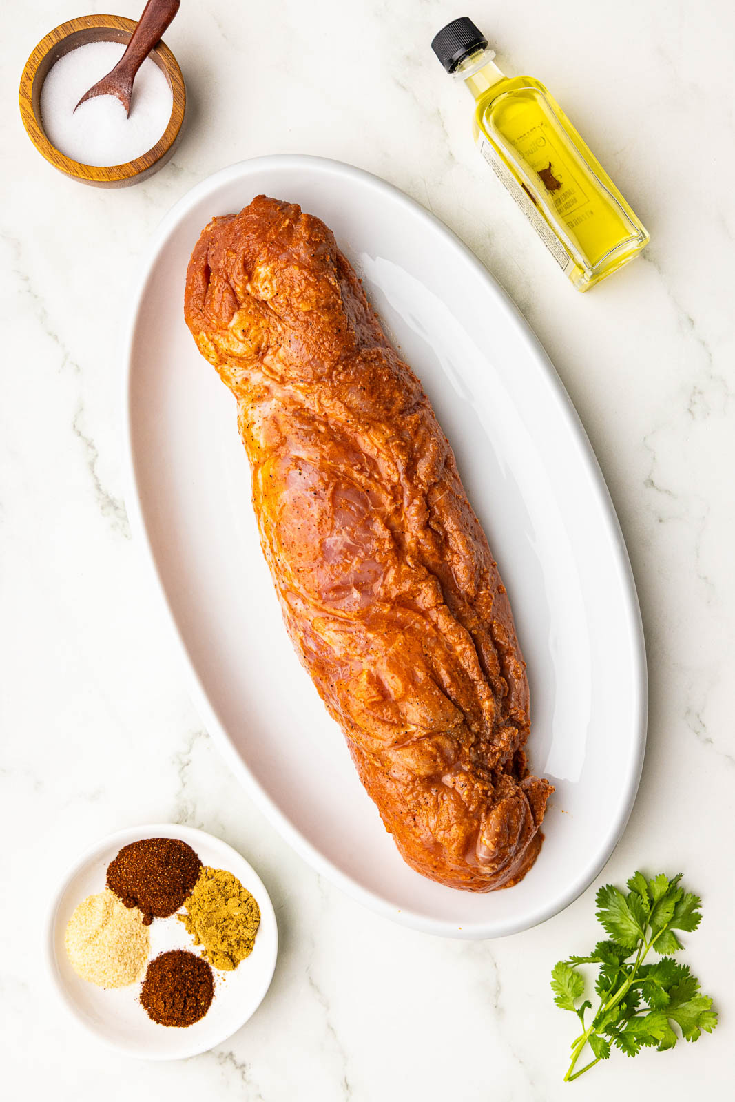 white platter containing pork tenderloin, surrounded by wooden bowl of salt bottle of olive oil, and small bowl of seasonings