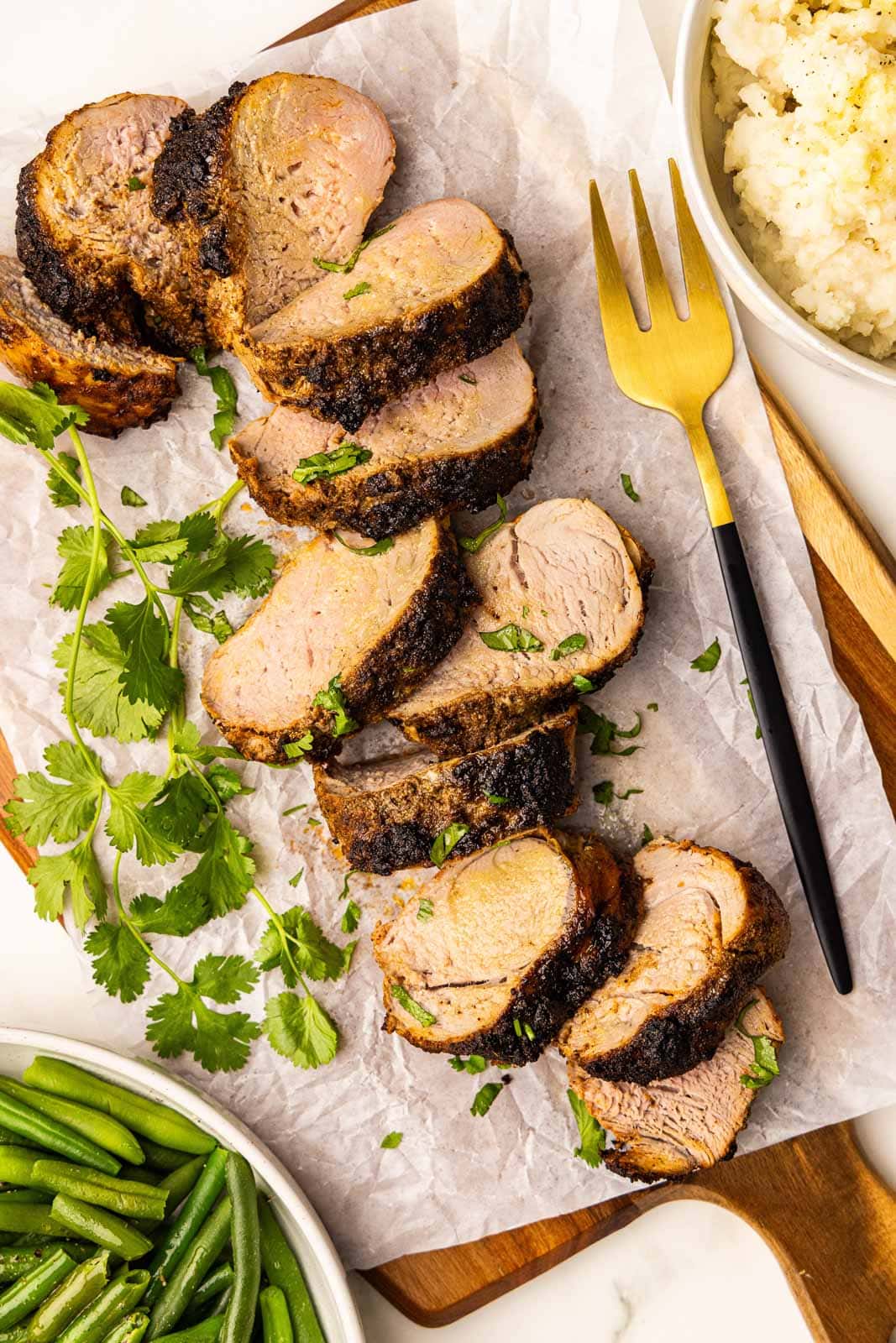 overhead view of sliced pork loin on parchment lined wooden cutting board with a gold fork, surrounded by bowls of green beans and mashed potatoes