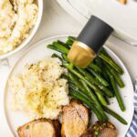 overhead view of white dinner plate of 4 pork loin slices, mashed potatoes and green beans, surrounded by bowl of potatoes and air fryer