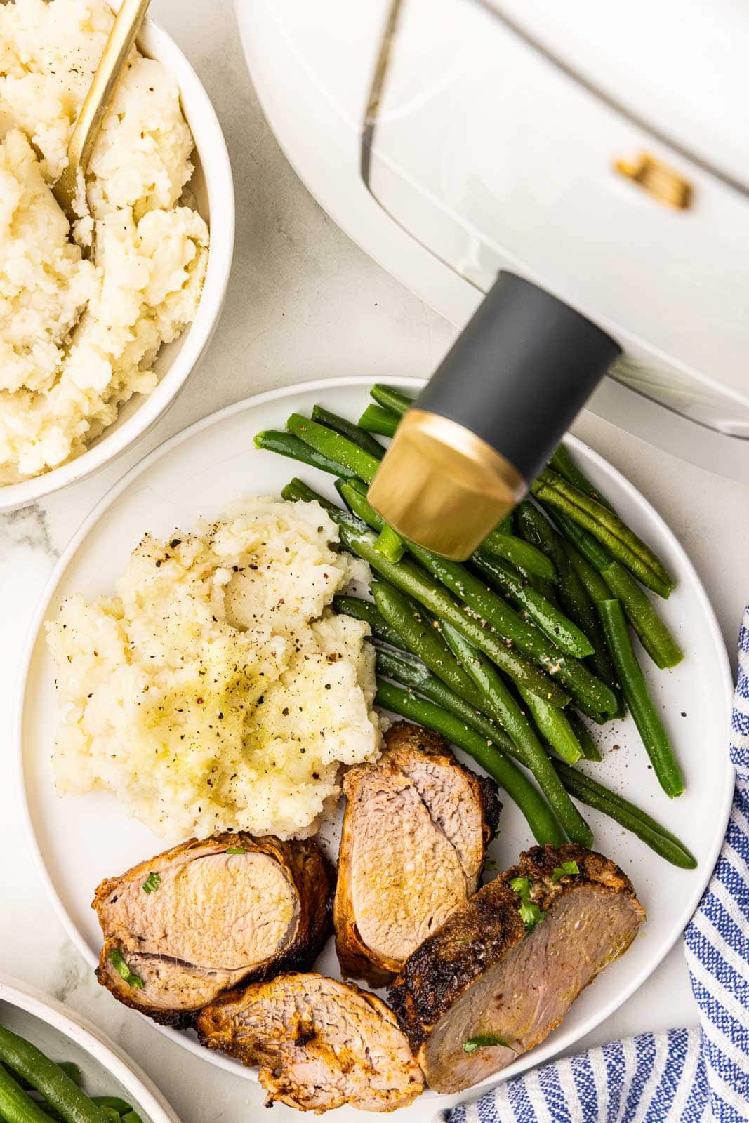 overhead view of white dinner plate of 4 pork loin slices, mashed potatoes and green beans, surrounded by bowl of potatoes and air fryer