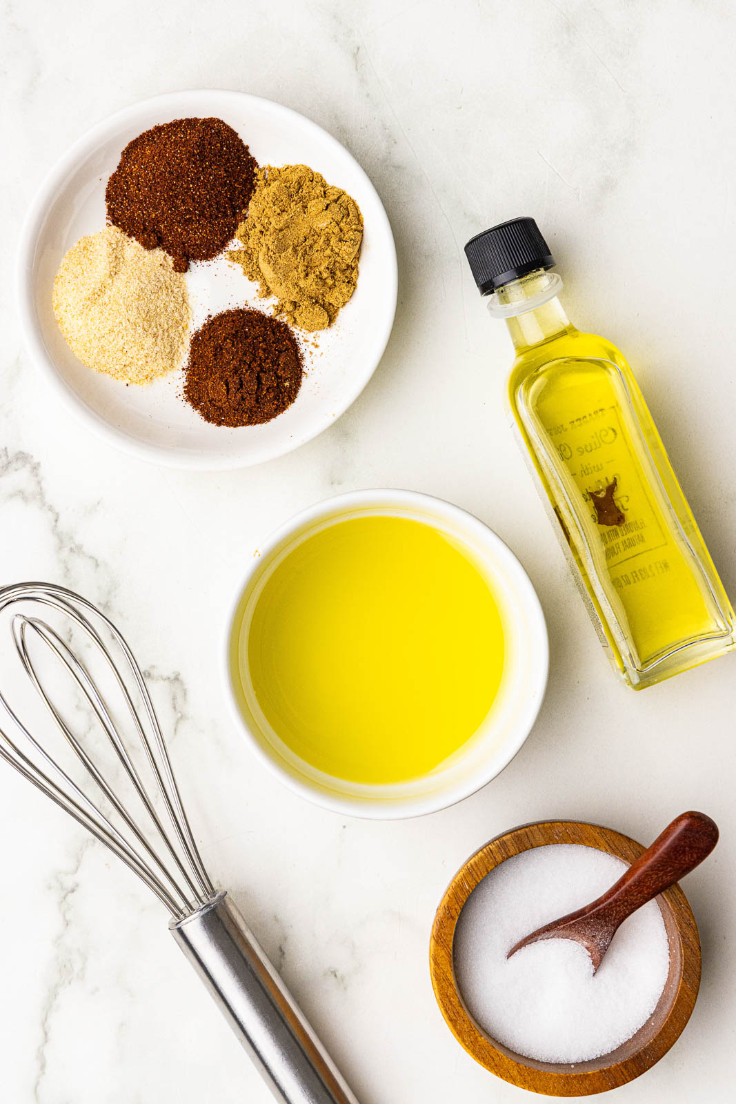 bowls of ingredients including seasonings, olive oil, bottle of olive oil, wooden bowl of salt and whisk