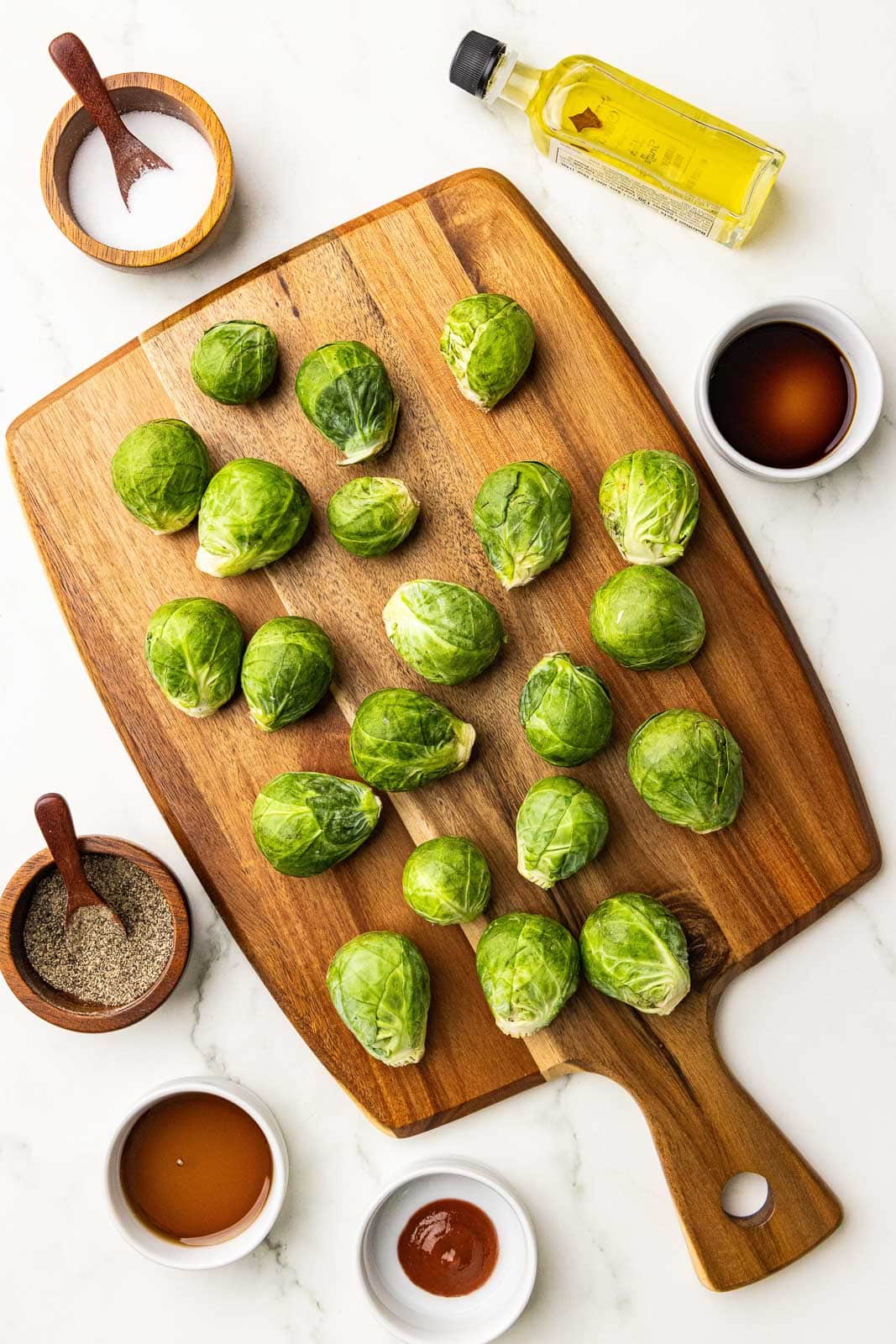 ingredients to include wooden cutting board with Brussels sprouts, surrounded by wooden bowls with spoons of salt and pepper, white bowls of maple syrup, Sriracha and soy sauce, and a bottle of olive oil
