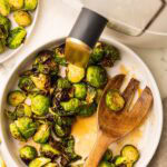 overhead view of white bowl of Brussels sprouts with glaze with a large wooden fork, with air fryer and another plate of Brussels sprouts in the background