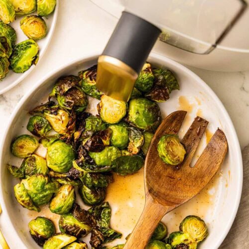 overhead view of white bowl of Brussels sprouts with glaze with a large wooden fork, with air fryer and another plate of Brussels sprouts in the background