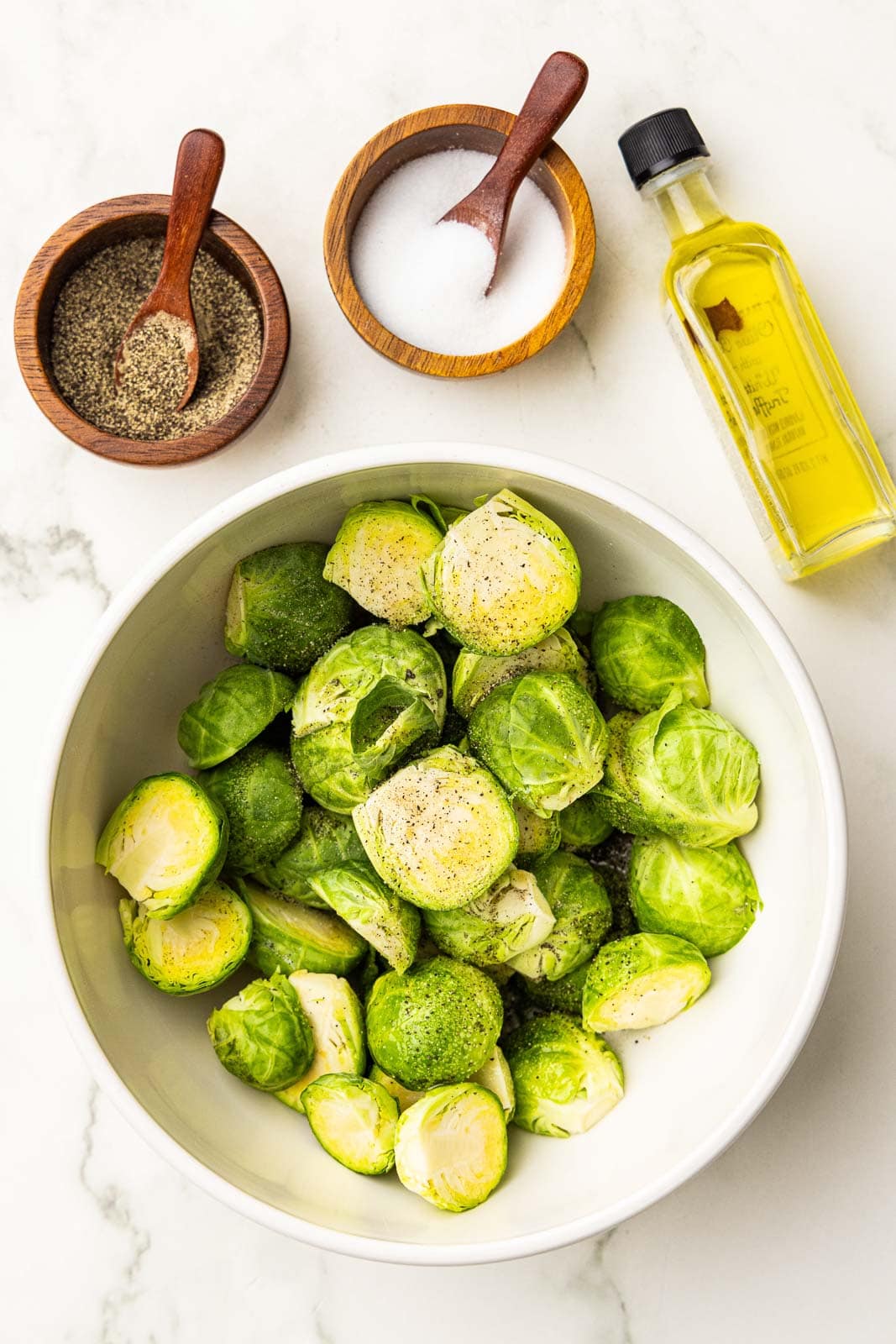 white bowl of prepared Brussels sprouts, with wooden bowls with spoons of salt and pepper and bottle of olive oil