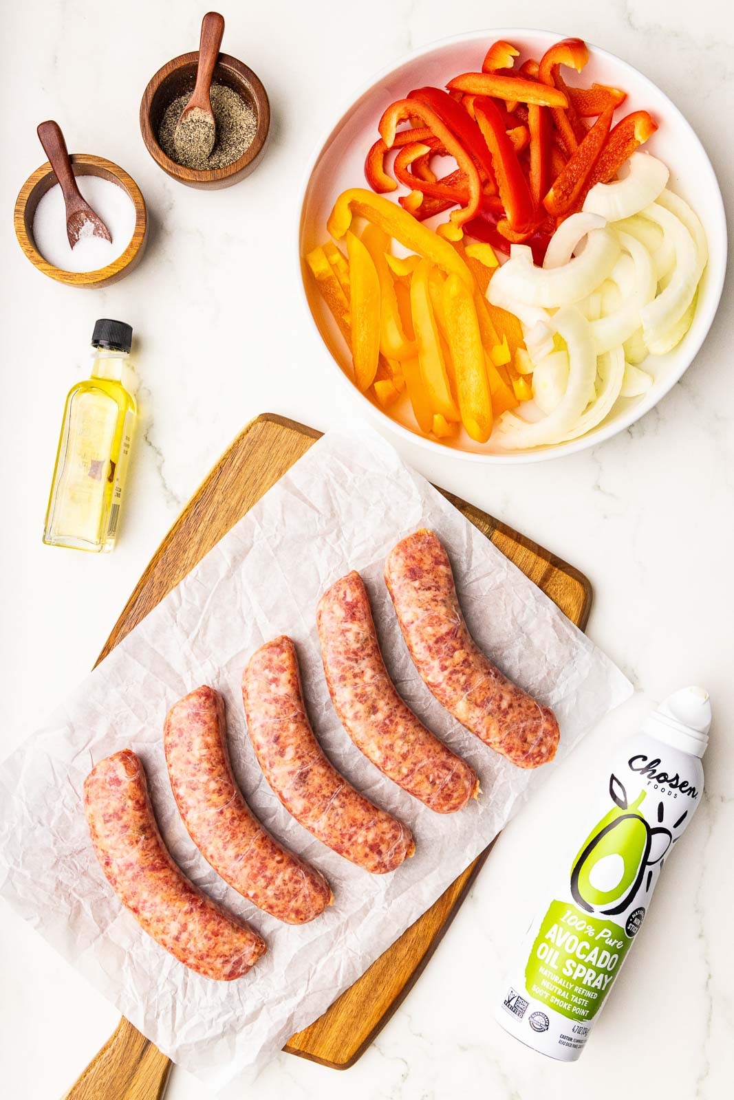 ingredients to include plate containing sliced red and yellow peppers and sliced, white onion, avocado oil spray, 5 sausage links on parchment lined wooden cutting board,