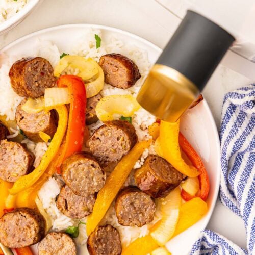 overhead view of white plate of cut up sausage with peppers, with white air fryer and bowl of rice in background