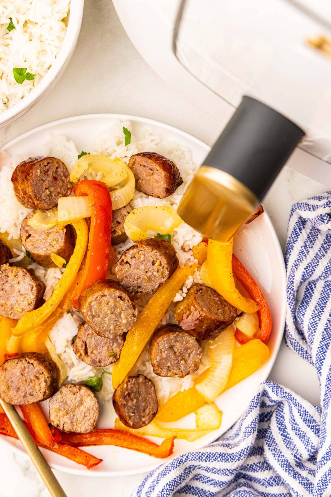 overhead view of white plate of cut up sausage with peppers, with white air fryer and bowl of rice in background