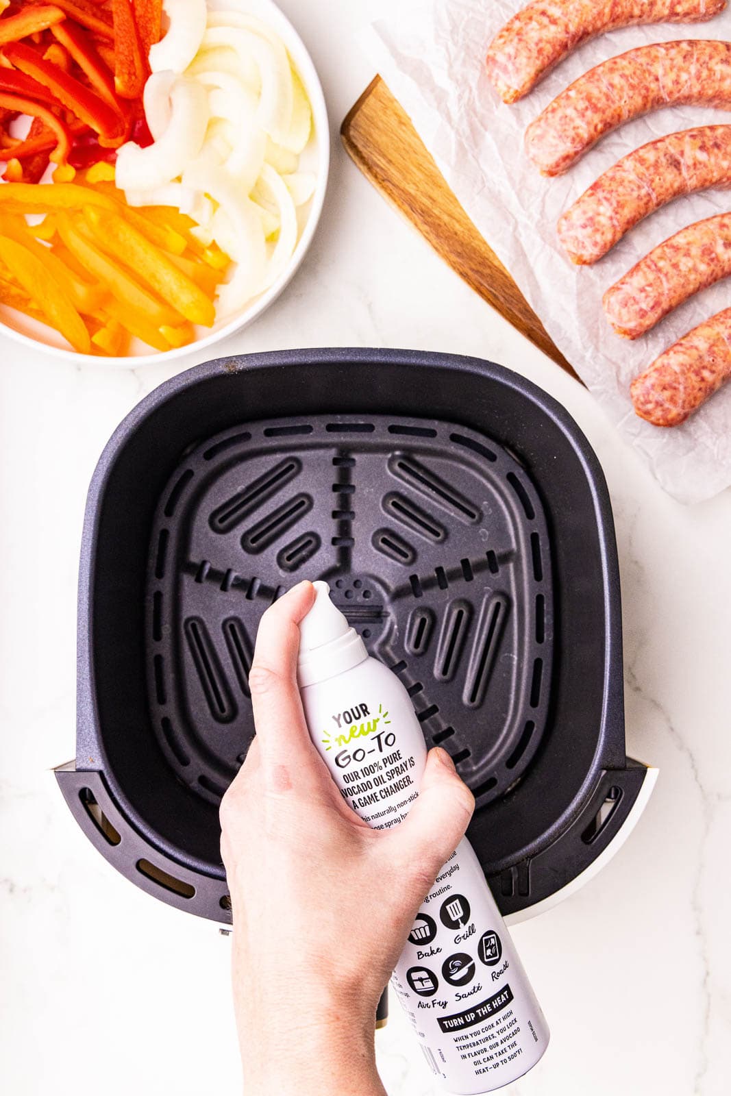 overhead view of black air fryer with hand spraying avocado spray, with plate of peppers and onions and sausages in background