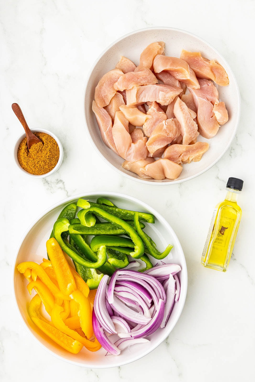 overhead view of white bowls containing cut up chicken breasts and sliced green and yellow peppers and purple onion, with small bowl of fajita seasoning and bottle of olive oil on side