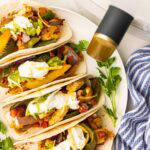 overhead view of four prepared fajitas in soft tortillas surrounded by a bowl of guacamole, white air fryer and blue and white striped dishtowel