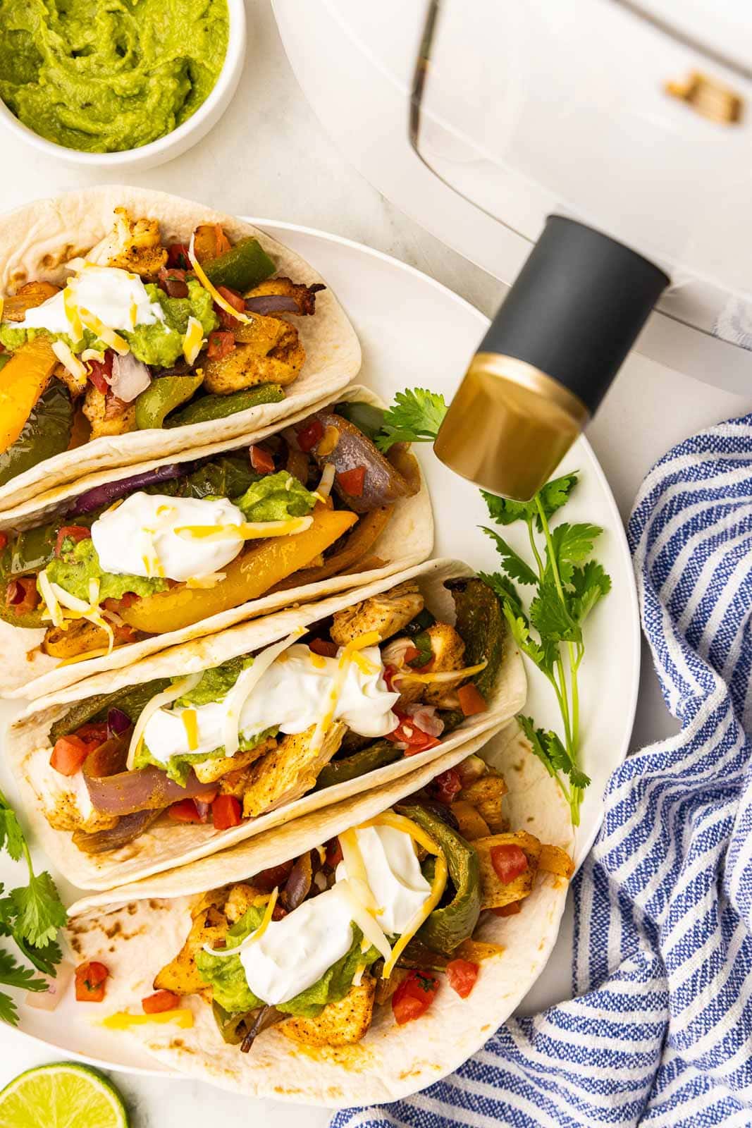 overhead view of four prepared fajitas in soft tortillas surrounded by a bowl of guacamole, white air fryer and blue and white striped dishtowel