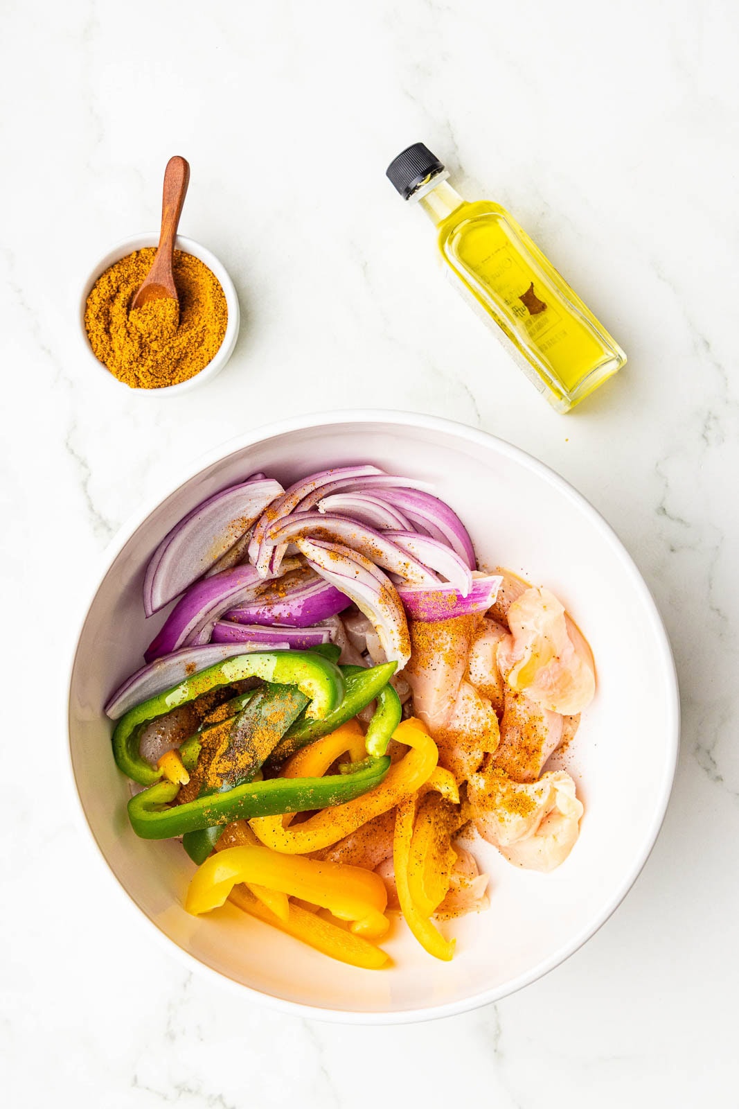 bowl of seasoned cut up chicken breasts, peppers and onions surrounded by bowl of fajita seasoning and bottle of olive oil