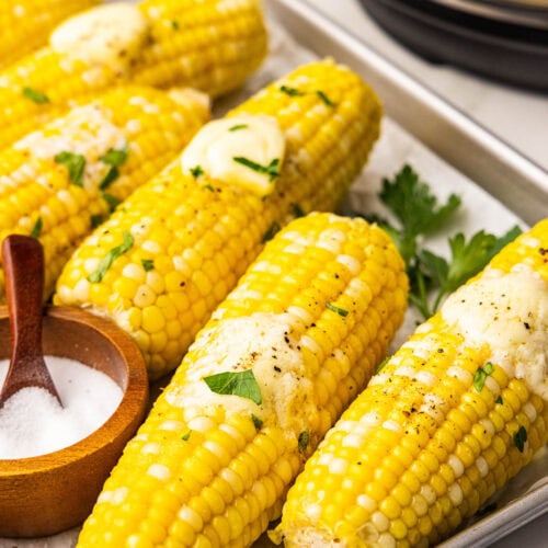 overhead side view of parchment lined cooking sheet containing six ears of corn on the cob, each with a pat of butter and wooden bowl with spoon of salt, with instant pot in background