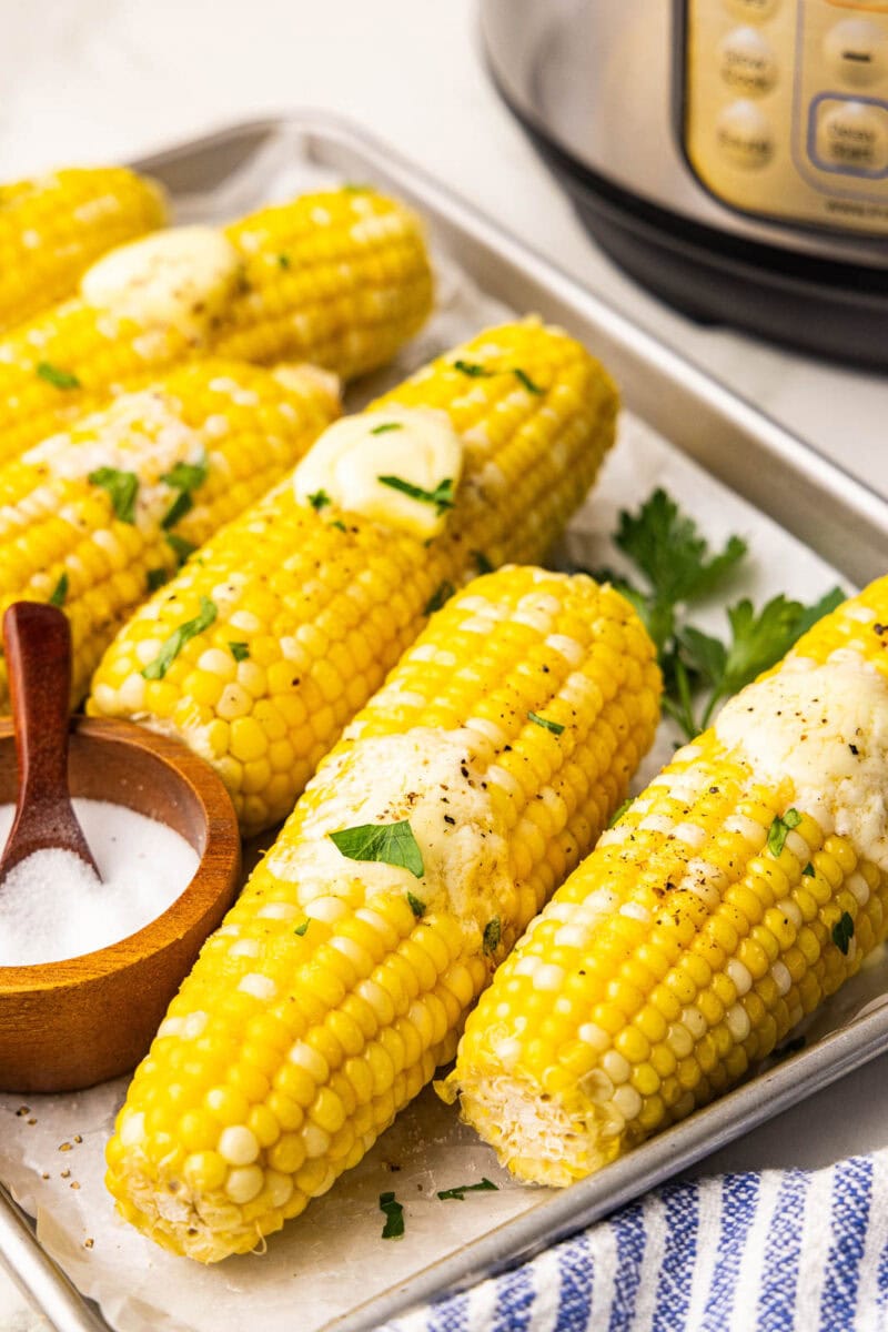 overhead side view of parchment lined cooking sheet containing six ears of corn on the cob, each with a pat of butter and wooden bowl with spoon of salt, with instant pot in background