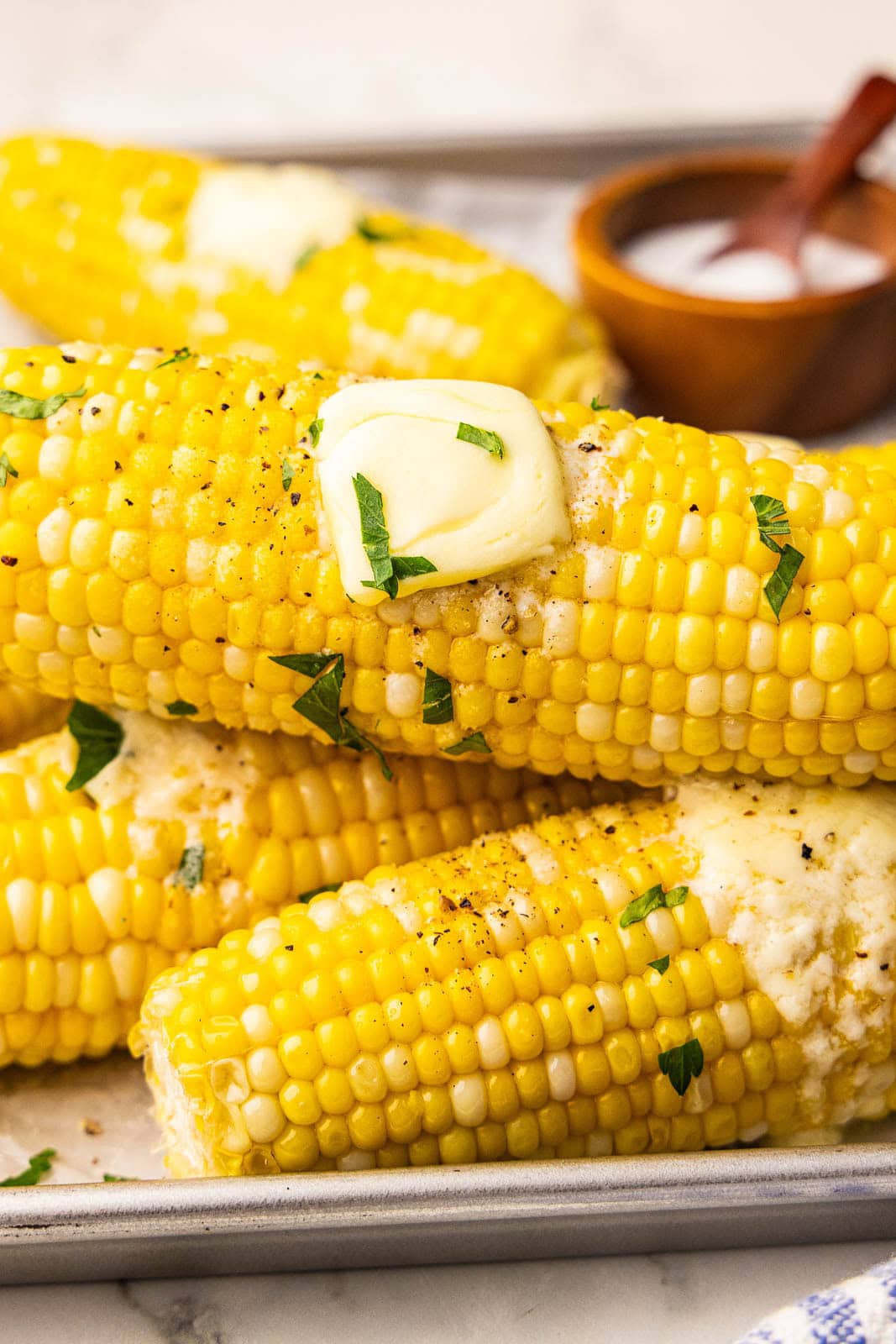 closeup side view of ears of corn on the cob with pats of butter with wooden bowl and spoon on salt in the background