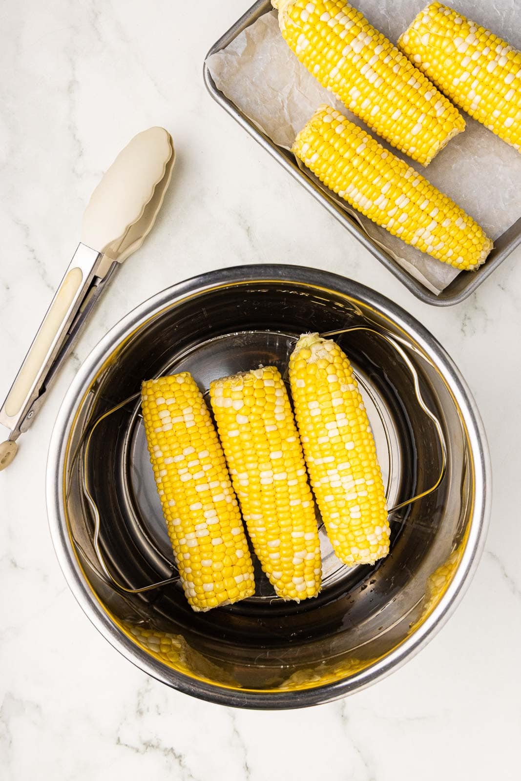 overhead view of instant pot containing 3 ears of corn with tongs and cooking sheet with 3 ears of corn on side